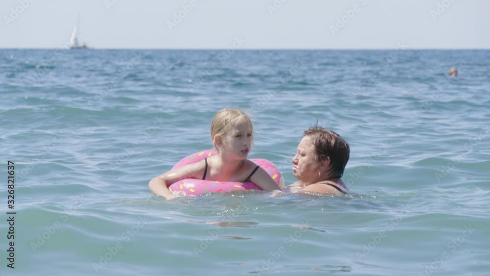 Grandmother with a small charming granddaughter bathe in the sea, happy family. Grandmother teaching little granddaughter to swim. Family resting on the beach. Grandmother with a small charming granddaughter bathe in the sea, happy family. Grandmother teaching little granddaughter to swim. Family resting on the beach.