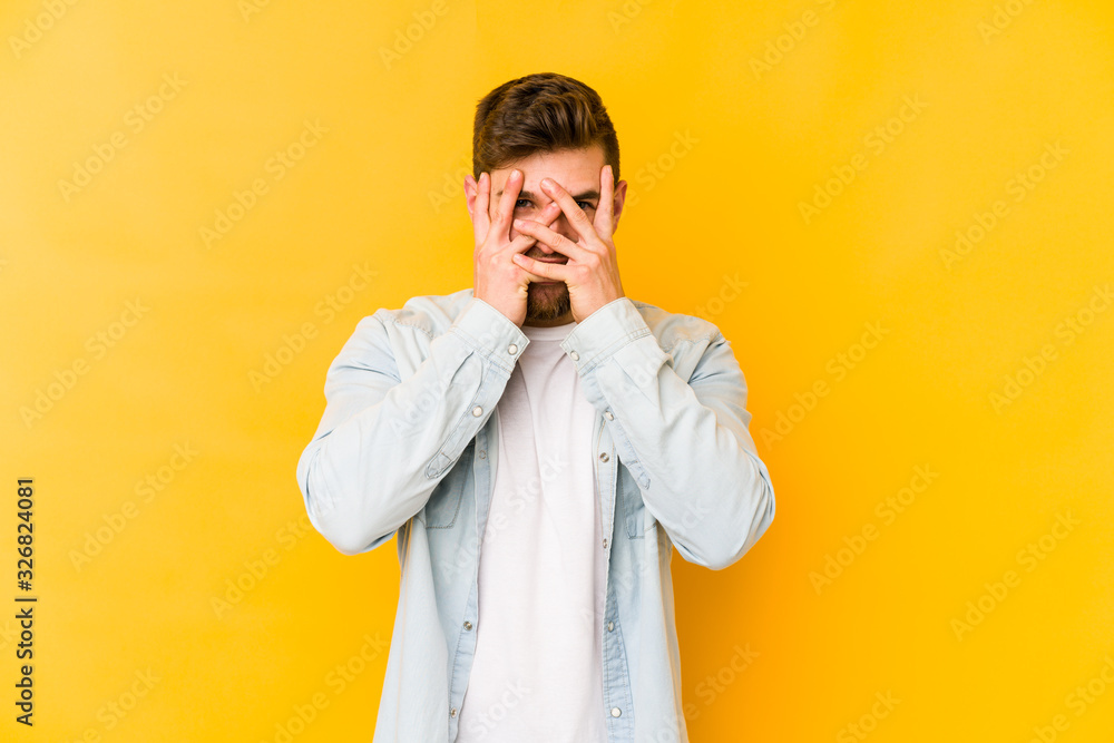 Young caucasian man isolated on yellow background blink at the camera through fingers, embarrassed covering face. Young caucasian man isolated on yellow background blink at the camera through fingers, embarrassed covering face.