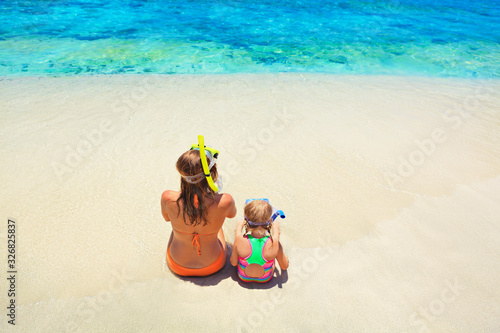 Fototapeta Naklejka Na Ścianę i Meble -  Happy family - mother, baby girl in snorkeling mask sit on white sand beach of tropical island. Look at scenic sea lagoon view. Travel adventure, swimming activity on summer holiday cruise with kids.
