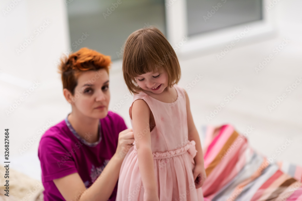 young mother helping daughter while putting on a dress