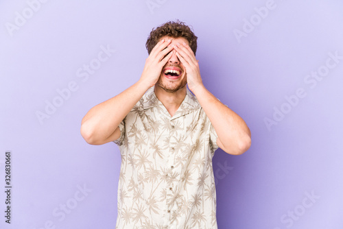 Young blond curly hair caucasian man isolated covers eyes with hands, smiles broadly waiting for a surprise.