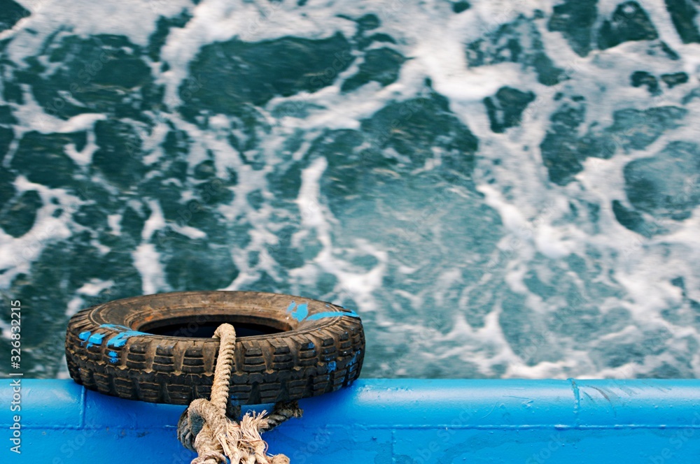 Fototapeta premium Rubber tire using as bumper protect side of boat or ship, bumper hanging on industrial boat for sea, top view and focusing at tire, waving sea water background, transportation concept