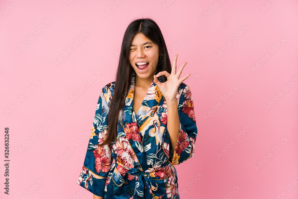 Young asian woman wearing a kimono pajamas winks an eye and holds an okay gesture with hand. Young asian woman wearing a kimono pajamas winks an eye and holds an okay gesture with hand.