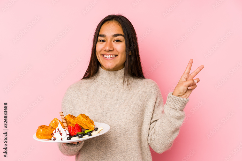Young asian woman holding a waffle isolated joyful and carefree showing a peace symbol with fingers. Young asian woman holding a waffle isolated joyful and carefree showing a peace symbol with fingers.