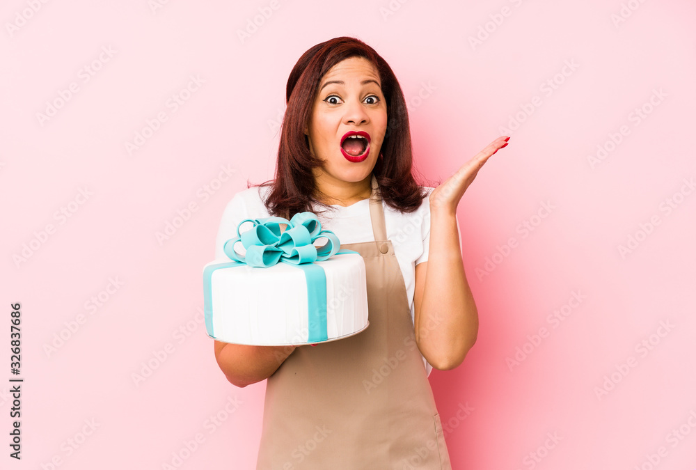 Middle age latin woman holding a cake isolated on a pink background surprised and shocked. Middle age latin woman holding a cake isolated on a pink background surprised and shocked.