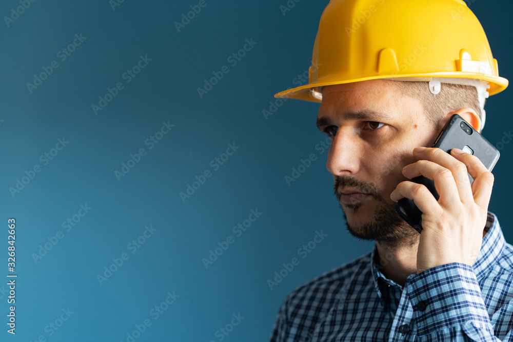 Close up portrait of young man engineer wearing yellow protecting safety helmet and shirt in front of blue wall isolated holding smart phone making a call talking side view Close up portrait of young man engineer wearing yellow protecting safety helmet and shirt in front of blue wall isolated holding smart phone making a call talking side view