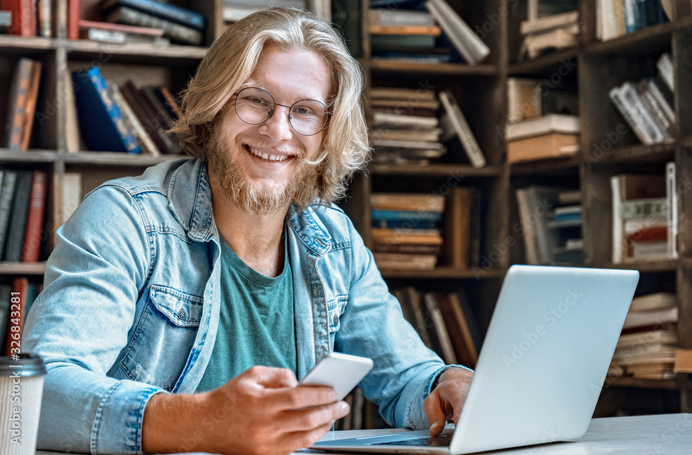 Portrait happy young man student hipster office home library desk hold modern smartphone use internet distant work laptop pc notebook send email study online smile look at camera education concept. Portrait happy young man student hipster office home library desk hold modern smartphone use internet distant work laptop pc notebook send email study online smile look at camera education concept.