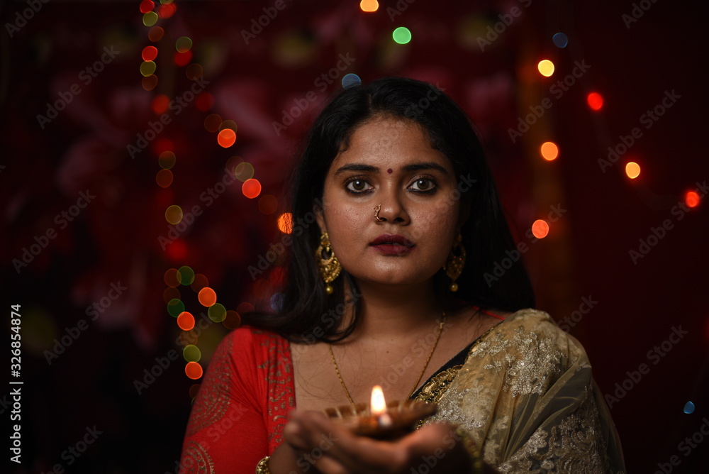 An young and beautiful Indian Bengali woman in Indian traditional dress is holding a Diwali diya/lamp in her hand standing in front of colorful bokeh lights. Indian lifestyle and Diwali celebration An young and beautiful Indian Bengali woman in Indian traditional dress is holding a Diwali diya/lamp in her hand standing in front of colorful bokeh lights. Indian lifestyle and Diwali celebration