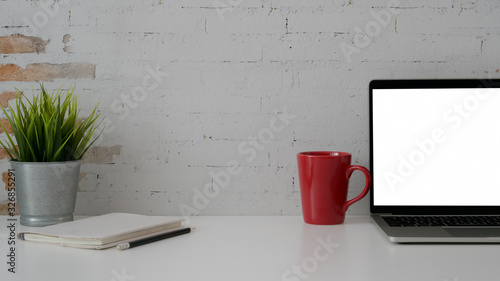 Close up view of workplace with blank screen laptop, stationery, red mug and tree pot on white table