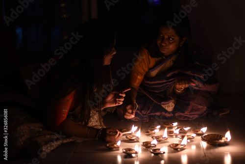 Two beautiful Indian Bengali women in Indian traditional dress are lighting Diwali diya/lamps sitting on the floor indoor in darkness on Diwali evening. Indian lifestyle and Diwali celebration