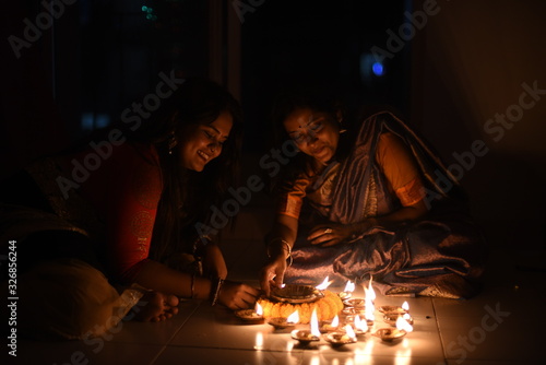 Two beautiful Indian Bengali women in Indian traditional dress are lighting Diwali diya/lamps sitting on the floor indoor in darkness on Diwali evening. Indian lifestyle and Diwali celebration