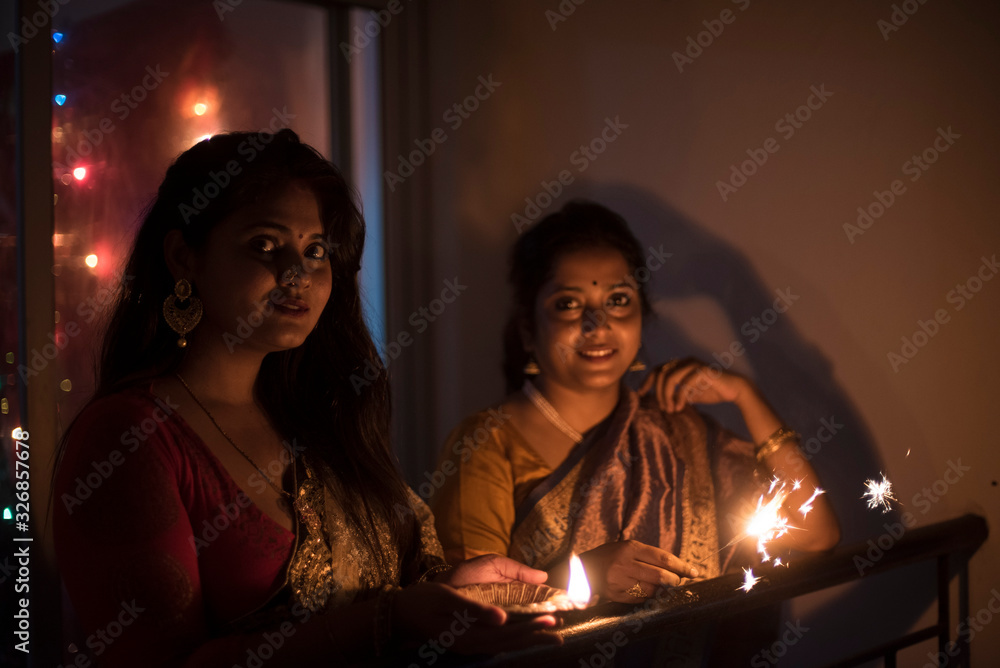 Two young and beautiful Indian Bengali women in Indian traditional dress are celebrating Diwali with diya/lamp and fire crackers on a balcony in darkness. Indian lifestyle and Diwali celebration Two young and beautiful Indian Bengali women in Indian traditional dress are celebrating Diwali with diya/lamp and fire crackers on a balcony in darkness. Indian lifestyle and Diwali celebration