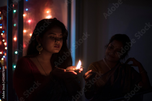 An young and beautiful Indian Bengali woman in Indian traditional dress is holding a Diwali diya/lamp in her hand standing on a balcony in darkness. Indian lifestyle and Diwali celebration