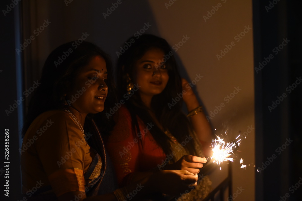 Two young and beautiful Indian Bengali women in Indian traditional dress are celebrating Diwali with diya/lamp and fire crackers on a balcony in darkness. Indian lifestyle and Diwali celebration Two young and beautiful Indian Bengali women in Indian traditional dress are celebrating Diwali with diya/lamp and fire crackers on a balcony in darkness. Indian lifestyle and Diwali celebration