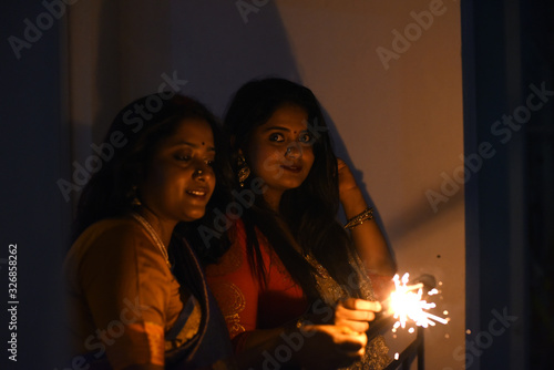 Two young and beautiful Indian Bengali women in Indian traditional dress are celebrating Diwali with diya lamp and fire crackers on a balcony in darkness. Indian lifestyle and Diwali celebration