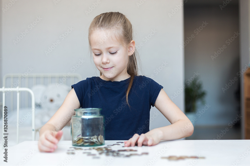 Cute little child girl counts and puts coins in a glass bottle in her room. The child saves money on the concept of the future Cute little child girl counts and puts coins in a glass bottle in her room. The child saves money on the concept of the future