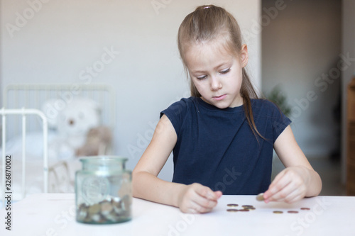 Cute little child girl counts and puts coins in a glass bottle in her room. The child saves money on the concept of the future