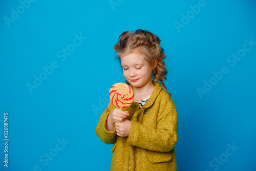 portrait of a baby girl in a coat holding a Lollipop on a colored blue background © Olesya Pogosskaya