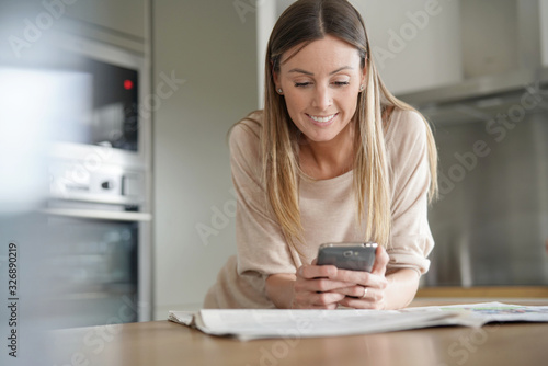 Woman in home kitchen reading newspaper and using smartphone