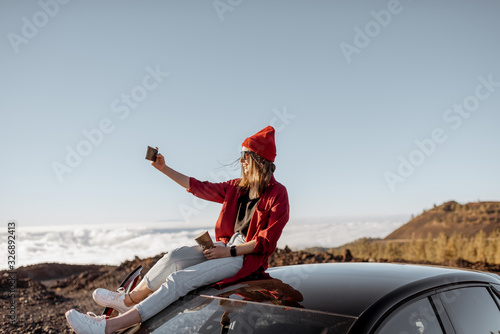 Young woman dressed in red enjoying rocky landscapes above the clouds, standing on the car highly in the mountains on a sunset. Carefree lifestyle and travel concept