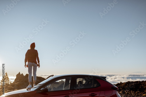Young woman dressed in red enjoying rocky landscapes above the clouds, standing on the car highly in the mountains on a sunset. Carefree lifestyle and travel concept. Image with copy space on the sky