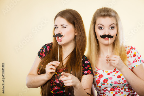 Two happy women holding fake moustache on stick