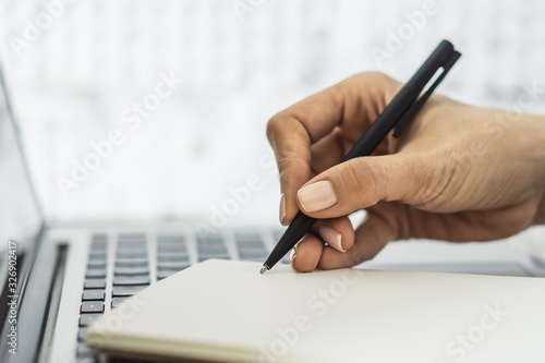 Woman writes with a pen in diary on laptop keyboard in a sunny office, business and education concept. Close up