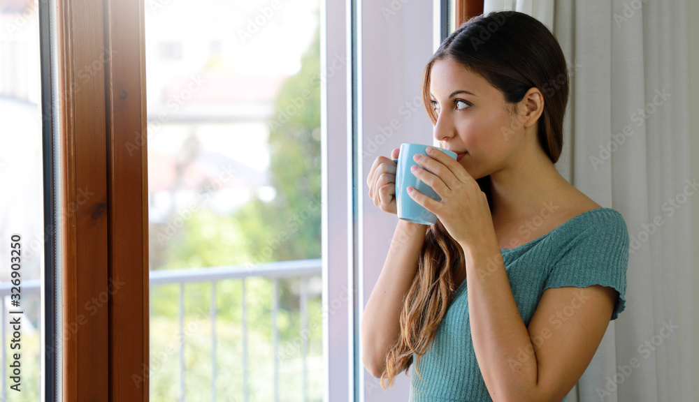 Portrait of beautiful woman looking through the window drinking cup of tea at home, focus on model eyes, indoor photo Portrait of beautiful woman looking through the window drinking cup of tea at home, focus on model eyes, indoor photo