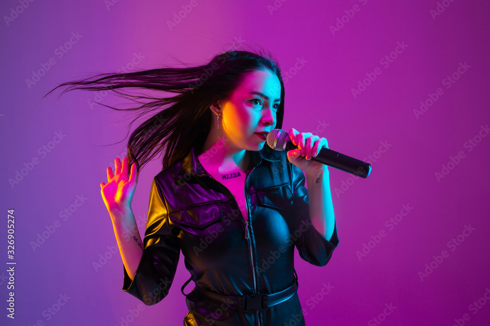 Caucasian female singer portrait isolated on purple studio background in neon light. Beautiful female model in black wear with microphone. Concept of human emotions, facial expression, ad, music, art. Caucasian female singer portrait isolated on purple studio background in neon light. Beautiful female model in black wear with microphone. Concept of human emotions, facial expression, ad, music, art.