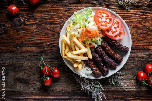 Fototapeta Naklejka Na Ścianę i Meble -  Spicy kofte (cevapcici, grilled minced meat) served with fresh tomatoes, lettuce, salad, vegetables and French fries on a rustic wooden board