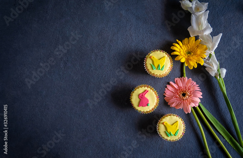 Pretty yellow vegan Easter cupcakes with colorful pastel icing shapes of Easter bunny rabbits, chicks together with Easter flowers