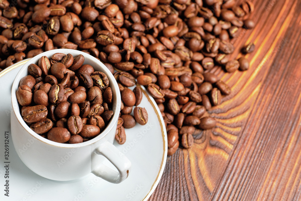 Coffee roasted grains are scattered in a white cup and on a wooden brown background. Coffee roasted grains are scattered in a white cup and on a wooden brown background.
