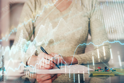 Hand taking notes in notepad. Forex chart holograms in front. Concept of research. Double exposure