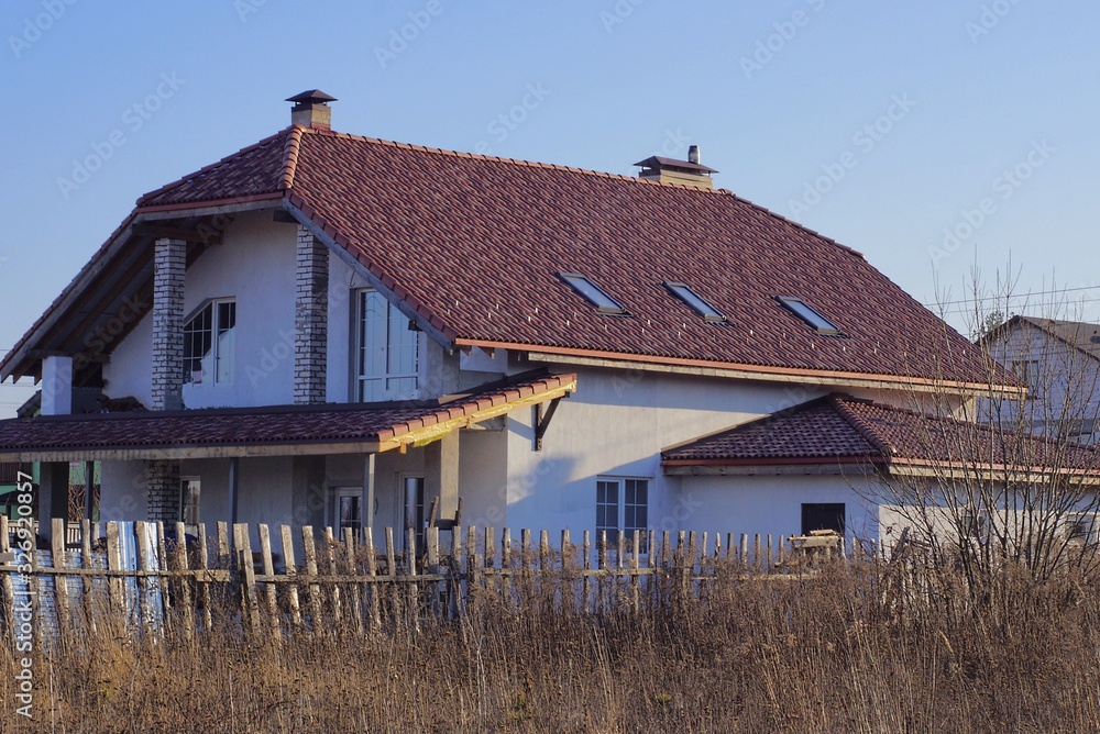 large white private house under a brown tiled roof with windows against a blue sky behind a wooden fence in dry gray grass large white private house under a brown tiled roof with windows against a blue sky behind a wooden fence in dry gray grass
