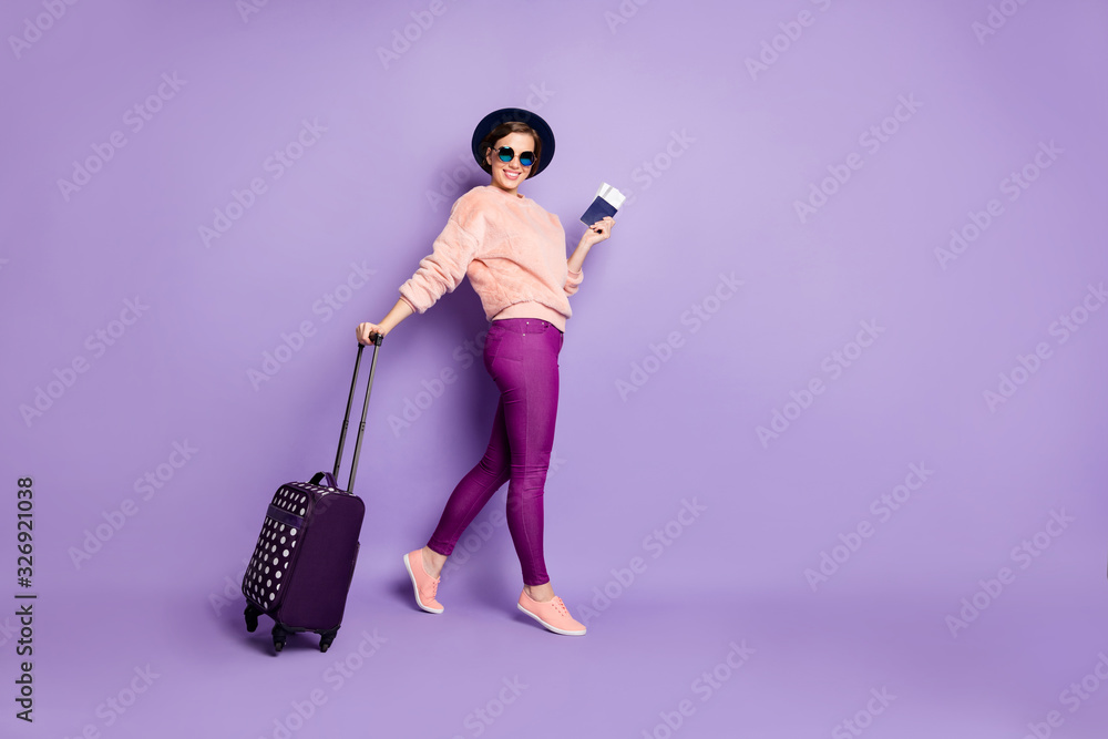 Full body photo of pretty lady vacation time walking registration flight hold ticket passport rolling bag wear hat sun specs casual pink sweater trousers isolated purple color background Full body photo of pretty lady vacation time walking registration flight hold ticket passport rolling bag wear hat sun specs casual pink sweater trousers isolated purple color background