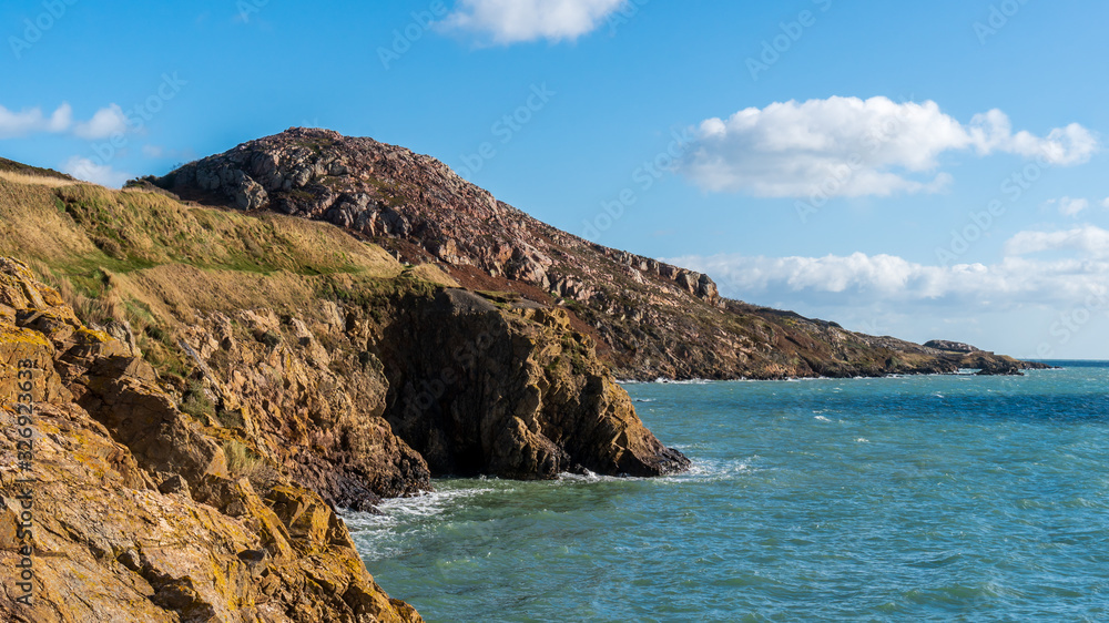 Fototapeta premium Irish landscape with rugged cliffs and blue sea. Part of the Howth Cliff Walk path with the Red Rock peak in the distance, near Sutton, Dublin, Ireland.