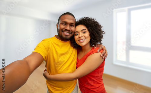 new home, real estate and people concept - happy smiling african american couple taking selfie over empty apartment on background