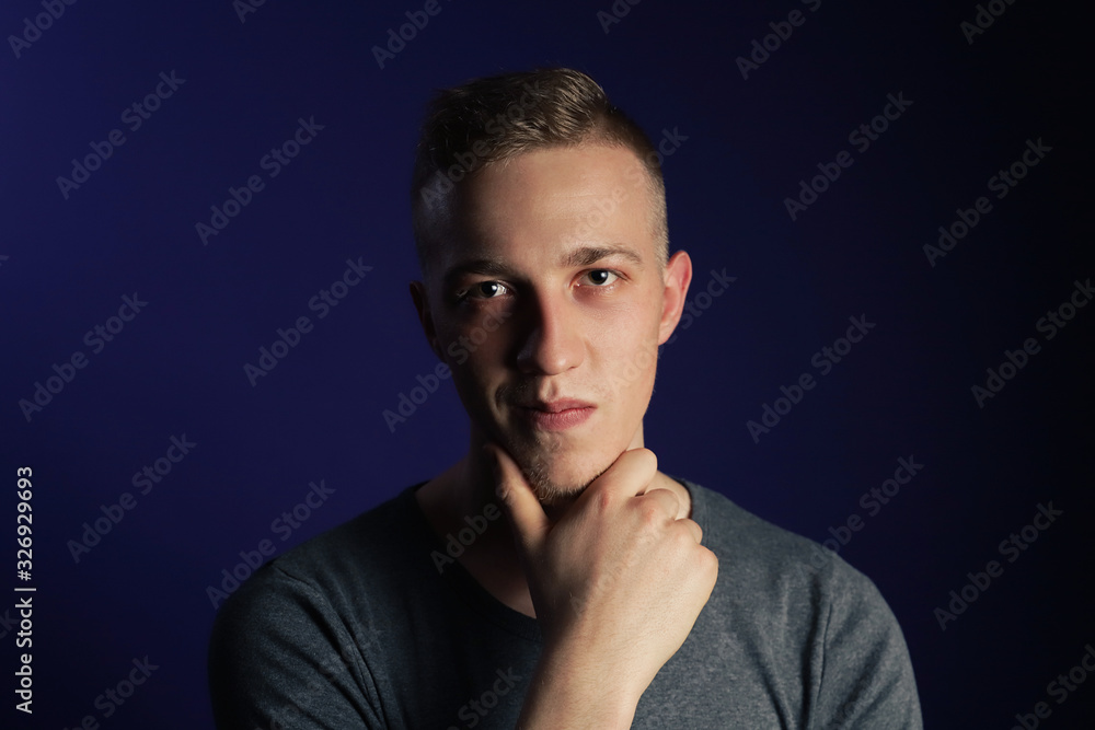 Young caucasian blonde man in grey t shirt on dark purple background touching his chin and thinking about something Young caucasian blonde man in grey t shirt on dark purple background touching his chin and thinking about something
