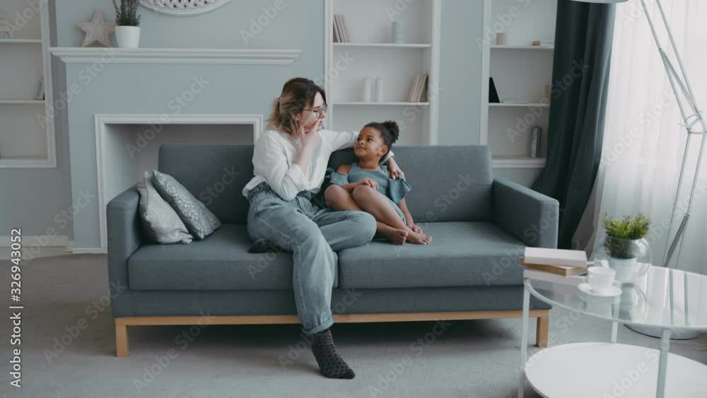 Beautiful young caring mother wearing shirt on sofa talking smart pretty african american daughter with curly black hair in blue dress, mum enjoying hugging spending time with little funny kid at home Beautiful young caring mother wearing shirt on sofa talking smart pretty african american daughter with curly black hair in blue dress, mum enjoying hugging spending time with little funny kid at home