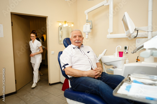 Portrait of happy senior man age 75 with mustache sitting in dentist   s chair  looking at camera while young dentist woman comes in the room to examine. Concept of dentistry  medicine and health care