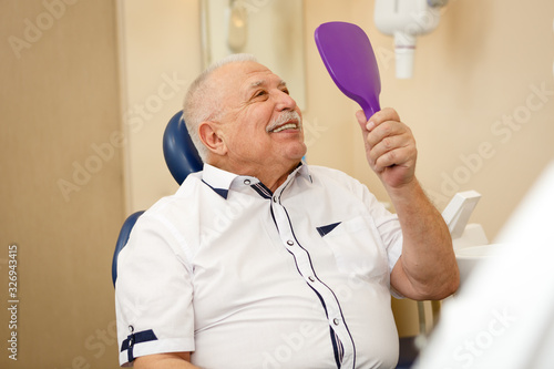Closeup portrait of happy satisfied senior man looking at mirror and enjoying his beautiful toothy smile after treatment. Dental care for old people. Concept of dentistry, medicine and health care.