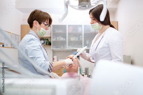 Modern stomatology private clinic with professional equipment. Two women dentists working with senior man patient. Dental care for elder people. Dentist holding dental device.