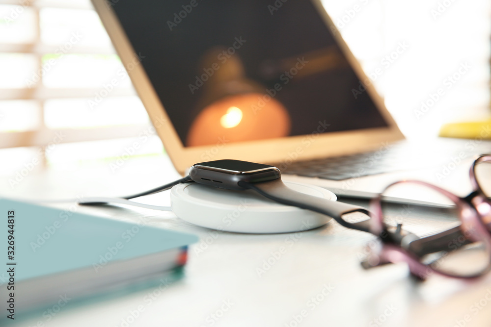 Obraz premium Smartwatch with wireless charger on white wooden table. Modern workplace accessory