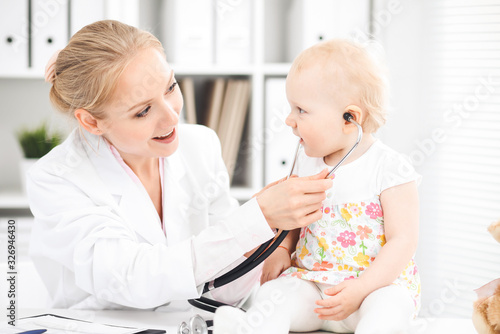 Doctor and patient toddler in hospital. Little girl dressed in dress with pink flowers is being examined by doctor with stethoscope. Medicine and health care concept