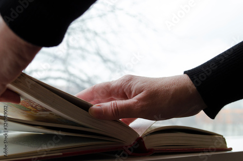 A person reading books near the window. Hands turns over book page.