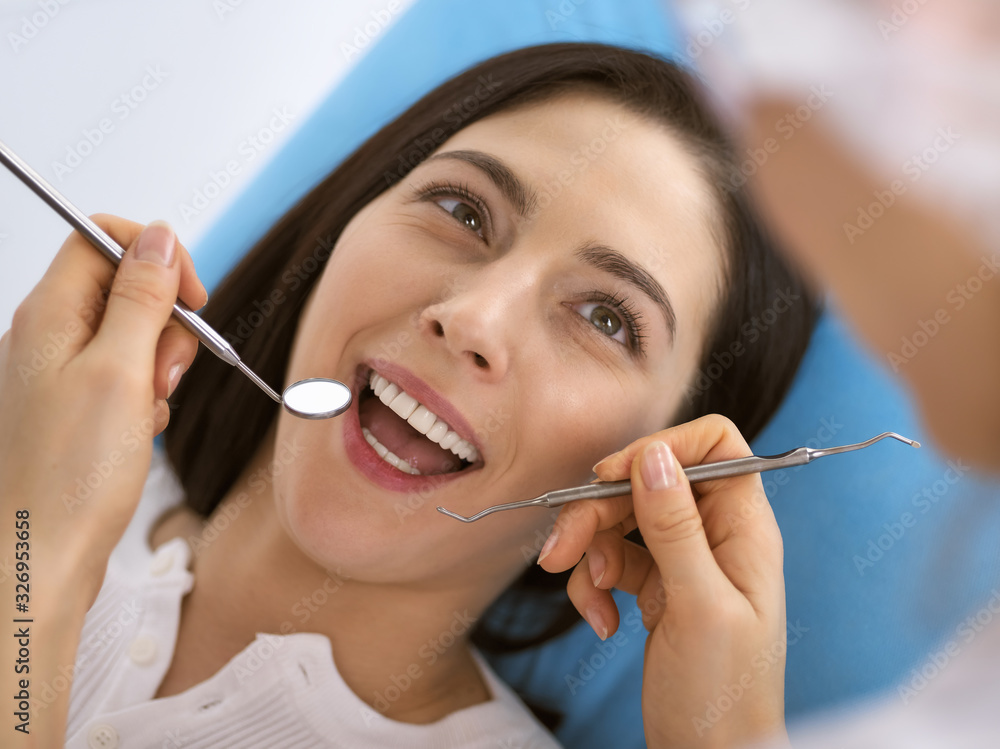 Smiling brunette woman being examined by dentist at dental clinic. Hands of a doctor holding dental instruments near patient's mouth. Healthy teeth and medicine concept