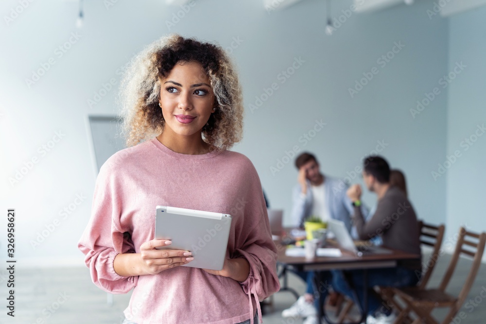 Fototapeta premium Business woman standing in foreground with a tablet in her hands, her co-workers discussing business matters in the background.