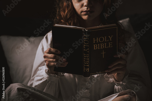 cropped view of creepy girl in nightgown reading bible on bed