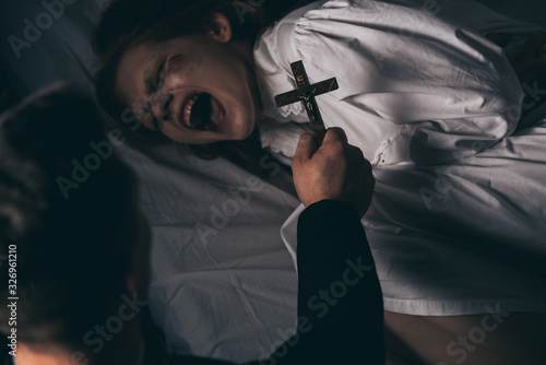 Bild auf Leinwand exorcist holding cross over demonic yelling girl in bed