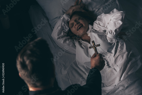 exorcist holding cross over demonic screaming girl in bed